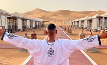 Alt: Campamento de lujo en el desierto de Merzouga con tiendas tradicionales, alfombra roja y faroles iluminados al atardecer en las dunas del Sahara.